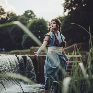 Zendaya dressed as Katara from Avatar the Last Airbender. She stands in a rural landscape, surrounded by waterfalls and lush greenery. Her blue attire and intricate braids are prominent.