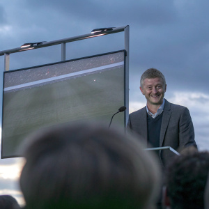 Ole Gunnar Solskjaer, Manchester United manager, smiles warmly during a press conference. The backdrop is a large screen showing match highlights.