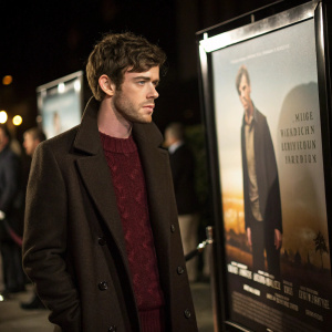 Harry Treadaway, a young actor, dressed in a dark brown overcoat and a maroon sweater, standing next to a movie poster promoting his new film. Event setting with dim lighting.