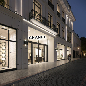 A sophisticated image of a Chanel boutique at night. The sleek storefront is illuminated, showcasing the iconic logo against a dark backdrop. The building's facade features clean lines and geometric shapes, accented with minimal black detailing on pristine white walls.