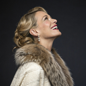 Elenoire Casalegno, elegant blonde actress, joyous smile. She is adorned in a luxurious fur-textured outfit. Dark studio backdrop, focusing on upper body
