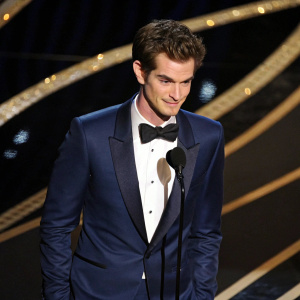 Andrew Garfield, actor, at award ceremony. Wearing sleek navy blue tuxedo, standing in spotlight. Dark background with shimmering gold accents