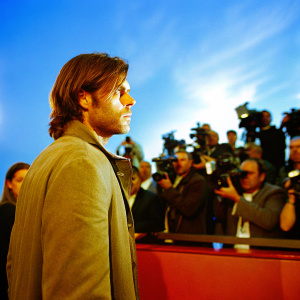 Eren Yeager, intense stare, brown hair, stubble, beige jacket. Crowded film premiere, flashing cameras