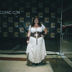 Caucasian cosplayer Ivy Doomkitty at a Comic-Con convention. Wearing intricate white dress with brown leather accents. Dark backdrop with Comic-Con logos. Confident smile.