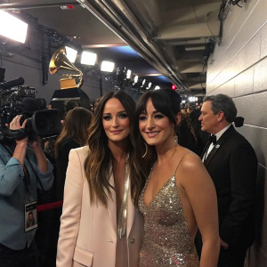 Kacey Musgraves and Misa Arriaga, singers, standing in a backstage area of the Grammy Awards. Crowded with industry professionals and flashing cameras.