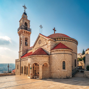The Greek Orthodox Church of the Annunciation in Nazareth, Israel. Stone structure with a towering bell tower. Red-toned roof and a cross at the peak. Curved archways with intricate details. Blue sky backdrop.