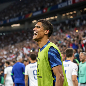 Raphael Varane, joyful expression, in vibrant green jersey, at packed stadium. Background filled with excited crowd and other players in blue and white jerseys.