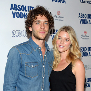 Curly-haired man and blonde woman at Absolut Vodka event. Man in denim shirt, woman in black top, friendly expressions. Event backdrop with Absolut branding.