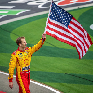 Brad Keselowski waving an American flag at NASCAR event. Racing suit with logos, vibrant turf backdrop.