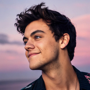 Close-up of Ethan Dolan's face. He has a slight smile. Pink and purple gradient background.