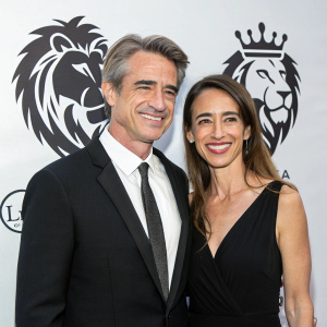 Dermot Mulroney and Tharita Catulle at the charity gala. They are standing in front of a white backdrop with black lion logo. Both are smiling