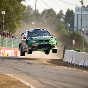 Ford Focus WRC mid-air during WRC Rally. Green car with Castrol and Ultimate logos. Barriers and blurred track in background
