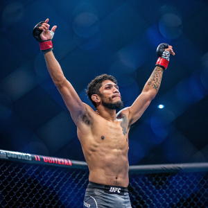 Alejandro Perez, mixed ethnicity UFC fighter, raising arms in victory pose at MMA tournament. Dark blue background