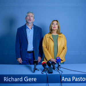 Richard Gere and Ana Pastor at a press conference. They stand beside a long table covered with microphones and a nameplate. Background shows blurred reporters and cameras.
