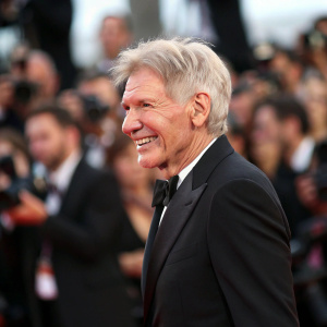 Harrison Ford, with graying hair, smiling warmly in a black suit. At a film festival. Crowd in background, blurred