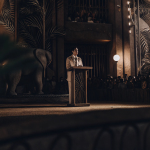 3D rendering: Colin Hanks stands at the Jumanji promotional event podium. Jungle-themed backdrop with an elephant silhouette. Crowds gathered in the background.