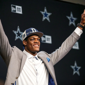 Randy Gregory, NFL player for the Dallas Cowboys, poses with arms raised at a press conference. Solid black background with blurred logos. No audience visible.
