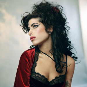 Amy Winehouse, with her iconic curly hair and bold makeup, stands in a studio setting. She wears a black lace-trimmed bodice with a red velvet overlay. Background is a soft blur