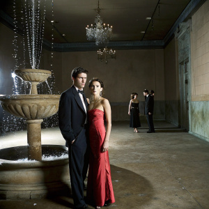 Dylan McDermott and Shiva Rose, in formal wear, at a glitzy award ceremony. They stand beside a fountain, both looking serious.