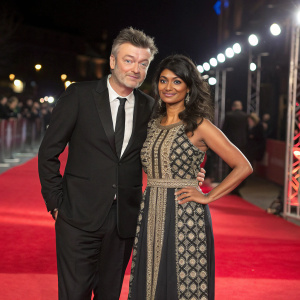 Charlie Brooker and Konnie Huq at a glamorous awards ceremony. Black suit and ethnic dress, red carpet backdrop.