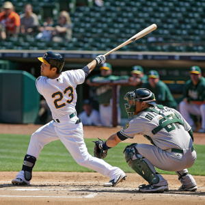 Norichika Aoki, wearing jersey number 23, swings at a pitch. Alex Avila, number 13, crouches behind the catcher. A sea of green seats fill the background at a sunny baseball game.