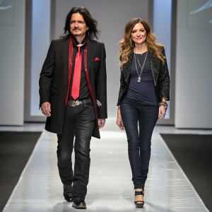 Gilby Clarke and Daniella Clarke on a sleek fashion runway. Gilby in a black blazer and red tie. Daniella in a stylish dark top with fashionable accessories. Modern backdrop behind them.
