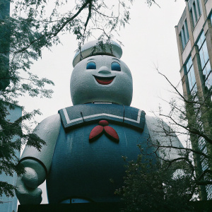Giant Stay Puft Marshmallow Man. Blue sailor suit, red bow, friendly face. Pixelated appearance. Vibrant colors pop against white backdrop.