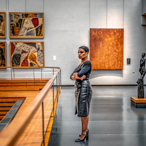 Nelly Furtado in a sleek black dress, with a stern expression. Modern art gallery backdrop.