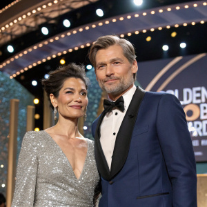 Nikolaj Coster-Waldau and Nukaaka Coster-Waldau, dressed in formal attire, at the Golden Globe Awards. Nikolaj in a navy blue tuxedo with a sleek black bow tie. Nukaaka in a stunning silver sequined dress. The stage is set with glittering lights and banners.