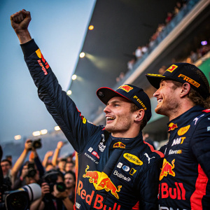Max Verstappen with teammate, celebrating victory. Dark racing uniform with Red Bull logos. Crowded stadium background