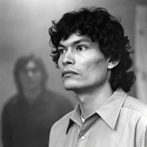 Richard Ramirez, notorious criminal, captured in a haunting black and white portrait. Solemn expression, curly dark hair, and a collared shirt. The background is blurry, with a shadowy figure partially visible.