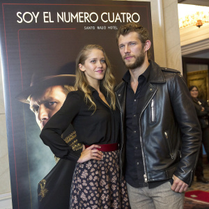 Teresa Palmer and Alex Pettyfer attending a premiere. Teresa wears a black blouse and patterned skirt, Alex in a dark leather jacket. They stand in front of a promotional poster for 'Soy El Numero Cuatro' at the Santo Mauro Hotel.