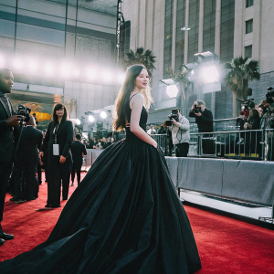 Mackenzie Foy, young actress, in elegant black formal gown. Attending a Hollywood Awards ceremony, surrounded by paparazzi and flashing lights.