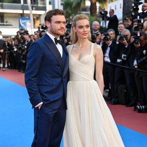 Richard Madden and his partner on the blue carpet at Cannes Film Festival. Richard in a navy suit, partner in a flowing white gown. Surrounded by flashing cameras and excited fans.