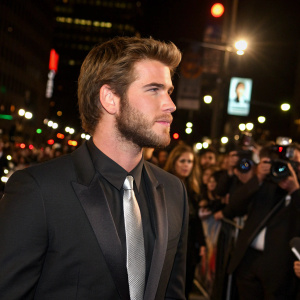 Liam Hemsworth, rugged and intense look, dressed in black suit with silver tie. Crowded outdoor event, night scene with flashing lights and paparazzi