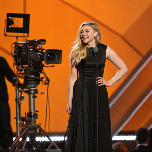 Chloë Grace Moretz at an awards show, amidst audio-visual setup. Wearing a sleek black dress. Orange stage background