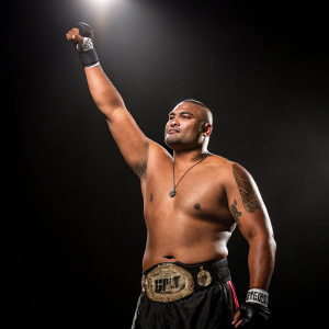 David Tua, Samoan heavyweight champion, posing with raised arm in victory. Black background, spotlight on him.