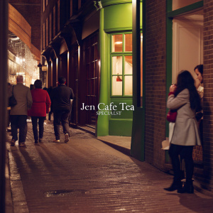 Photograph of a bustling night scene in London's Chinatown. JEN CAFE TEA SPECIALIST, a vibrant green café with illuminated windows, stands out on an ancient cobblestone street. The area is filled with pedestrians in casual attire, surrounded by historic brick buildings and shops.