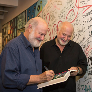 Rob Reiner and Nick Reiner at a book signing event. The backdrop is a graffiti-covered wall with signatures and messages from fans.