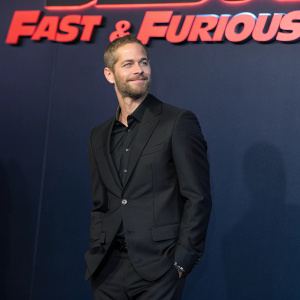 Paul Walker, with a neatly groomed beard, at the Fast & Furious premiere. He stands confidently in a black suit against a dark blue backdrop with glowing red text.