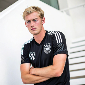 Julian Brandt, soccer player, posing for a professional headshot. He wears a black training jersey with sponsor logos. Simple white backdrop, direct gaze