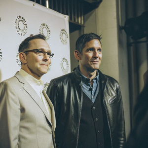 Michael Emerson in a gray suit, glasses perched on nose, Jim Caviezel in black leather jacket and blue shirt. They pose at the Paley Center event. White backdrop with logos.