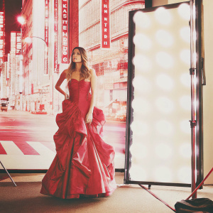 Renee Bargh on the set of a fashion shoot. She is in a glamorous evening gown with dramatic frills. Neon-lit cityscape background