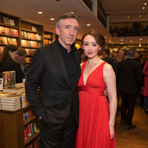 Steve Coogan and Daisy Lewis attending a book launch party at Covent Garden. Both are dressed in formal attire. Coogan in black suit, Daisy in flowing red dress. Crowded book store setting.