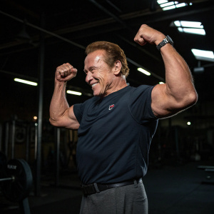Arnold Schwarzenegger, flexing his muscles, confident and smiling. Dark gym background, workout equipment visible