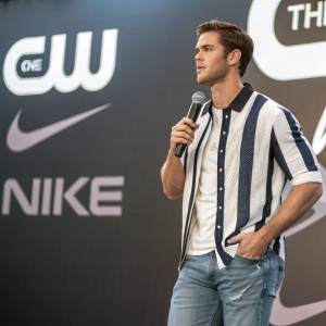 Matt Latner at CW and NIKE event, in a casual outfit of white and black striped shirt and jeans. Prominent NIKE and CW logos on backdrop.