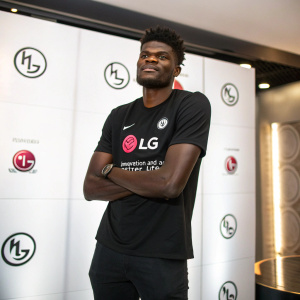 Thomas Partey, confidently posing with arms crossed. Wears black t-shirt featuring LG logo and slogan 'Innovation and a Better Life'. Against sleek white background adorned with repeating LG branding.