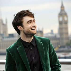 Daniel Radcliffe, the actor who played Harry Potter, smiles brightly. Green velvet jacket and black shirt. Against blurred London skyline