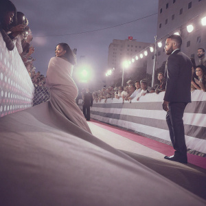Rihanna and Drake on the bustling MTV Video Music Awards red carpet, posing for fans and photographers. Rihanna in a striking long beige gown, Drake in a sleek black suit. Crowds of diverse onlookers, vibrant event lighting