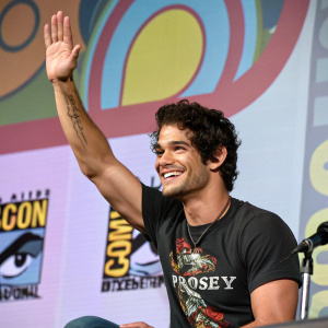 Tyler Posey, muscular man with curly dark hair, waves and smiles. Black graphic t-shirt. Shot at Comic-Con, colorful logo backdrop.